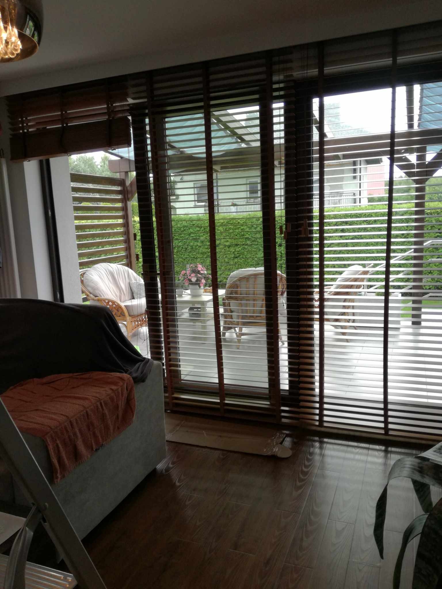 Drewniane żaluzje okienne zamontowane w domu w Woli Lubeckiej, widok z wnętrza na taras z meblami ogrodowymi i zielonym żywopłotem.