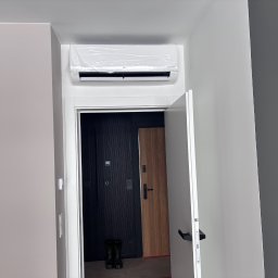 Air Klimat - Nowy klimatyzator LG, jeszcze w folii ochronnej, zamontowany nad drzwiami wejściowymi w nowoczesnym wnętrzu. Widoczne dwie pary butów przy drzwiach.