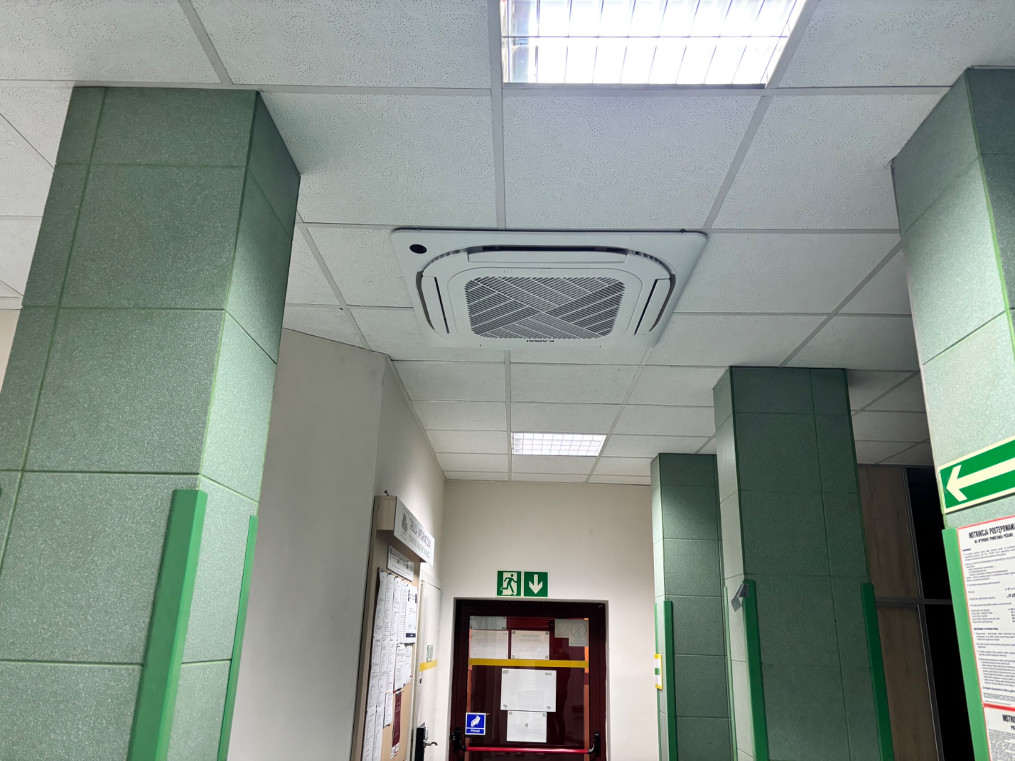 Kasetonowa jednostka klimatyzacji zamontowana w suficie podwieszanym biurowca, widok z dołu na tle korytarza z zielonymi kolumnami i drzwiami ewakuacyjnymi.