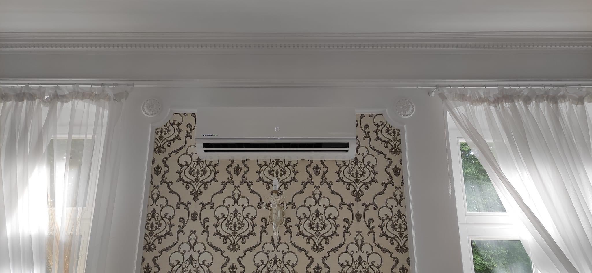 Biały klimatyzator Kaisai Eco zamontowany na ścianie z wzorzystą tapetą w eleganckim wnętrzu z białymi zasłonami i sztukaterią.
