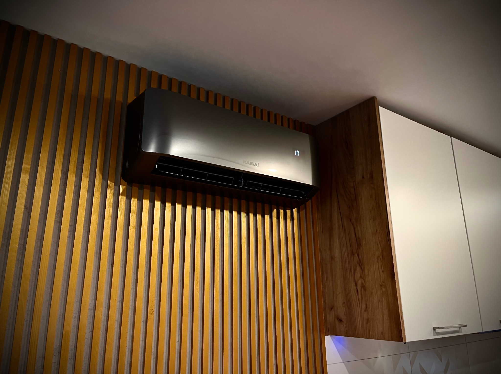 Szara jednostka klimatyzacyjna Kaisai zamontowana na ścianie z drewnianymi listwami w domu w Wałczu, widoczny wyświetlacz temperatury i fragment zabudowy kuchennej.
