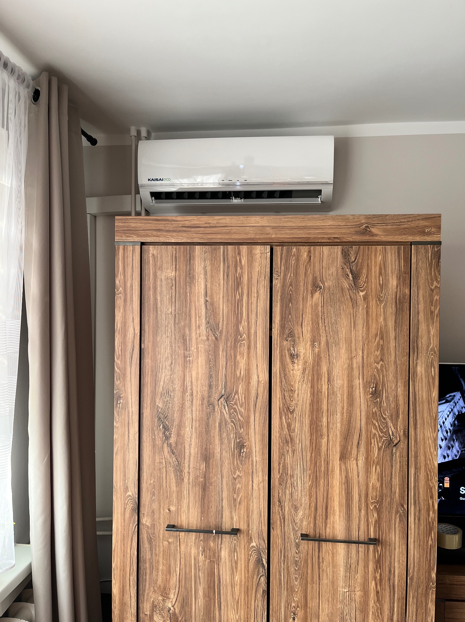 Biała jednostka klimatyzacji Kaisaieco zamontowana nad wysoką, dwudrzwiową szafą z drewnianym wzorem w jasnym pomieszczeniu.