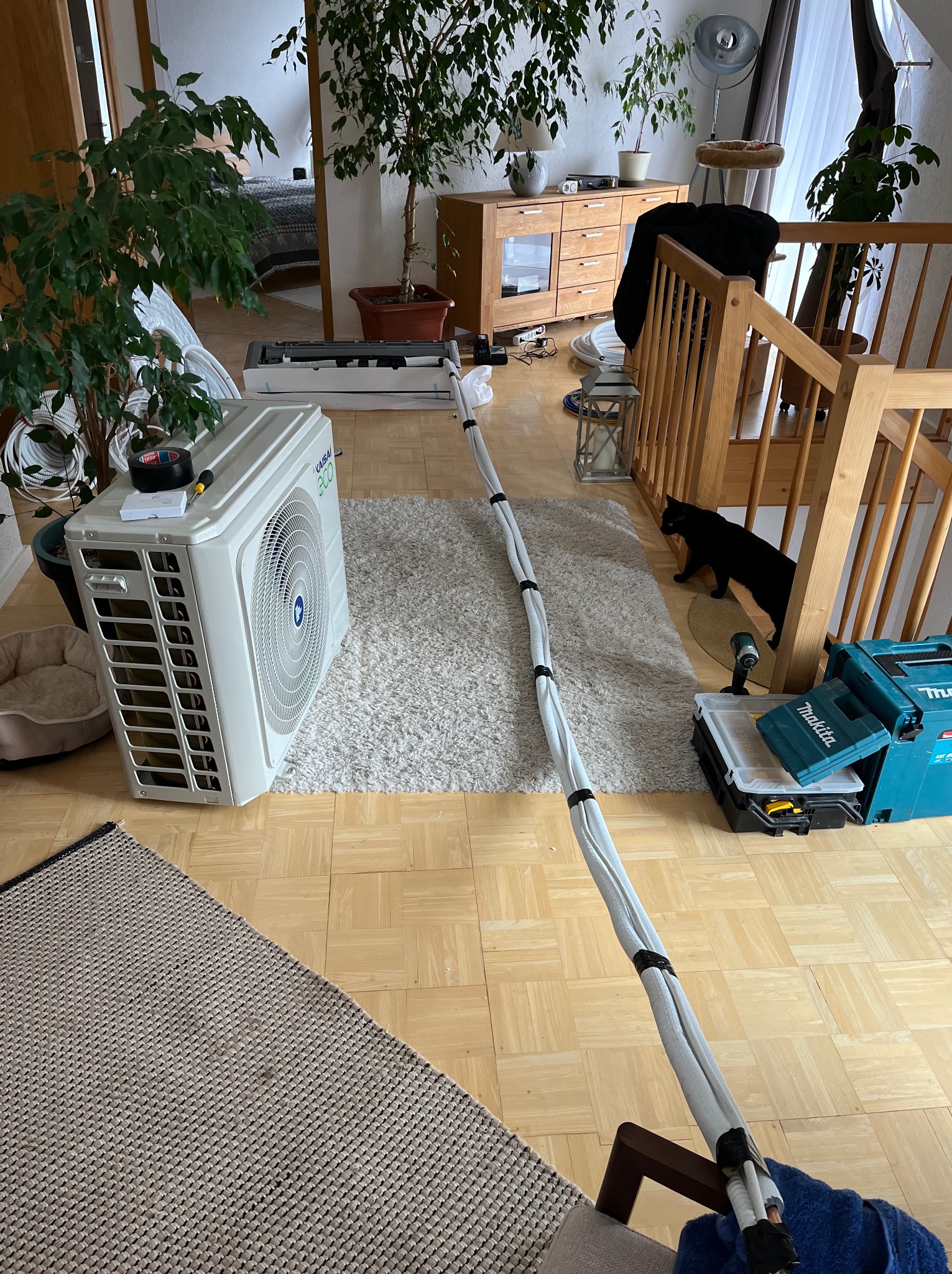 Wnętrze domu podczas instalacji klimatyzacji: jednostka wewnętrzna na dywanie, rury prowadzone po podłodze, skrzynka narzędziowa Makita i czarny kot przy schodach.