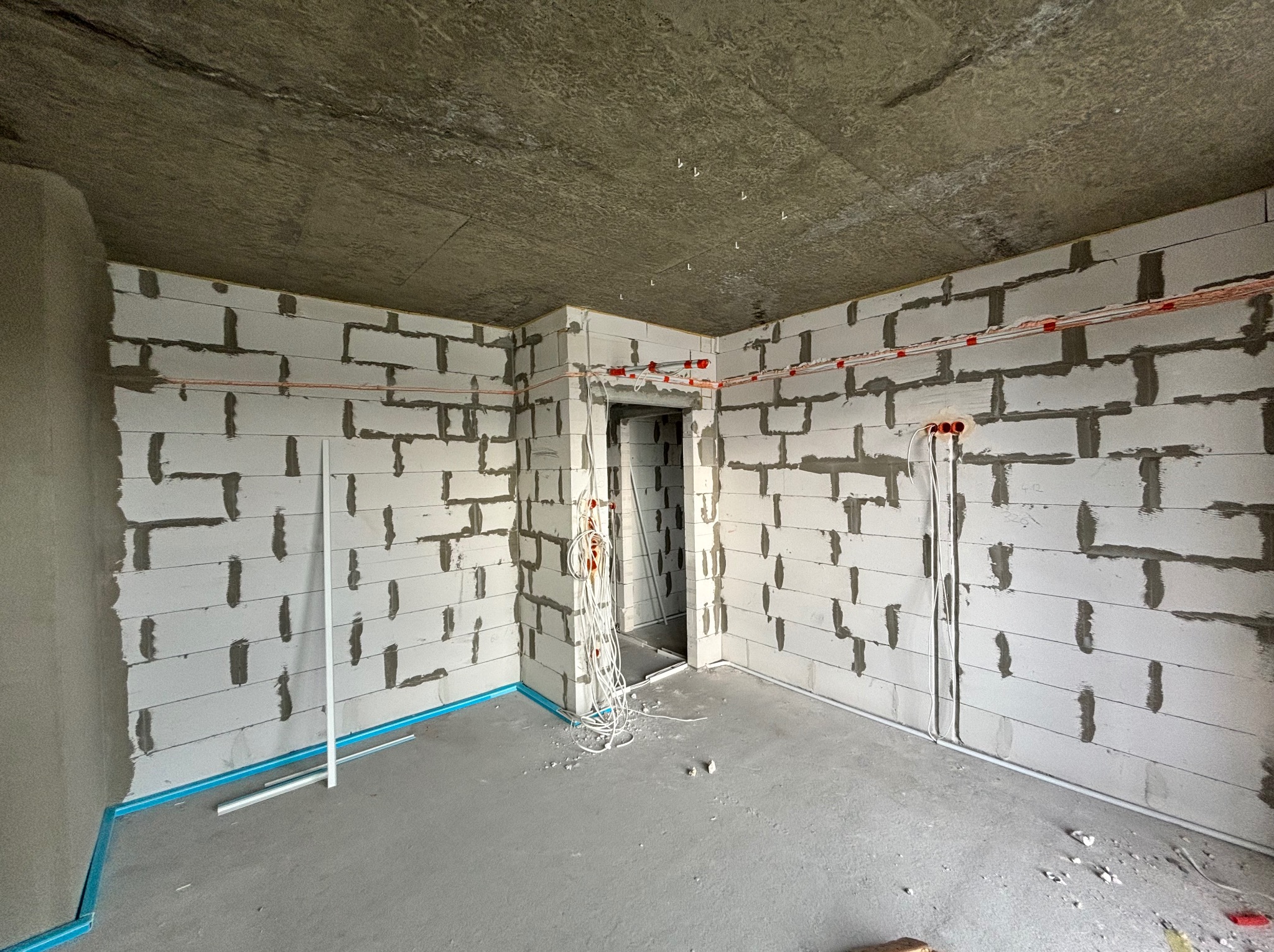 Surowy stan wnętrza w trakcie remontu: ściany z betonu komórkowego, widoczne przewody elektryczne, wylewka podłogowa, przygotowane do montażu rurki przy podłodze.