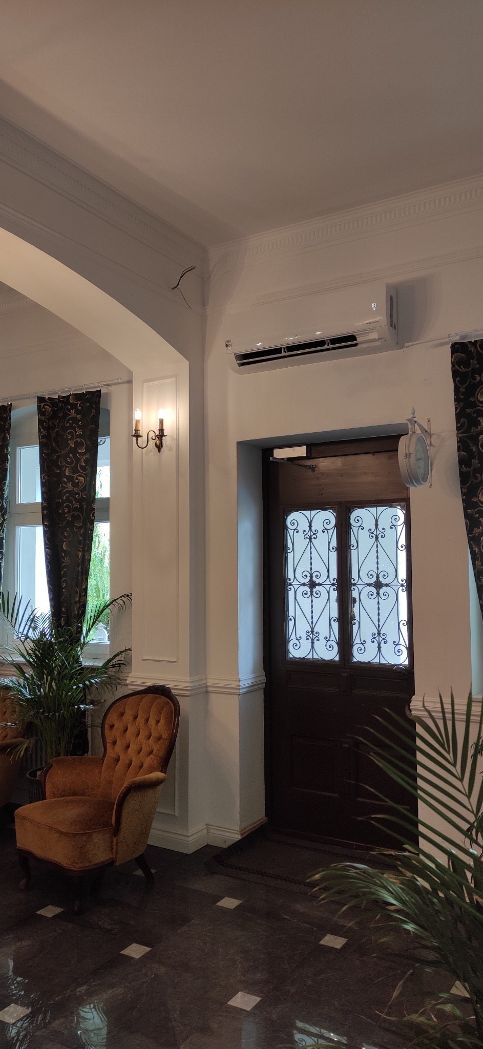 Biały klimatyzator LG zamontowany nad ciemnymi drzwiami z ozdobnymi kratami w eleganckim wnętrzu z roślinami doniczkowymi i fotelem w stylu vintage.