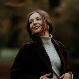 Uśmiechnięta kobieta w brązowym płaszczu i kremowym golfie, pozująca w jesiennym plenerze z rozmytym tłem drzew.