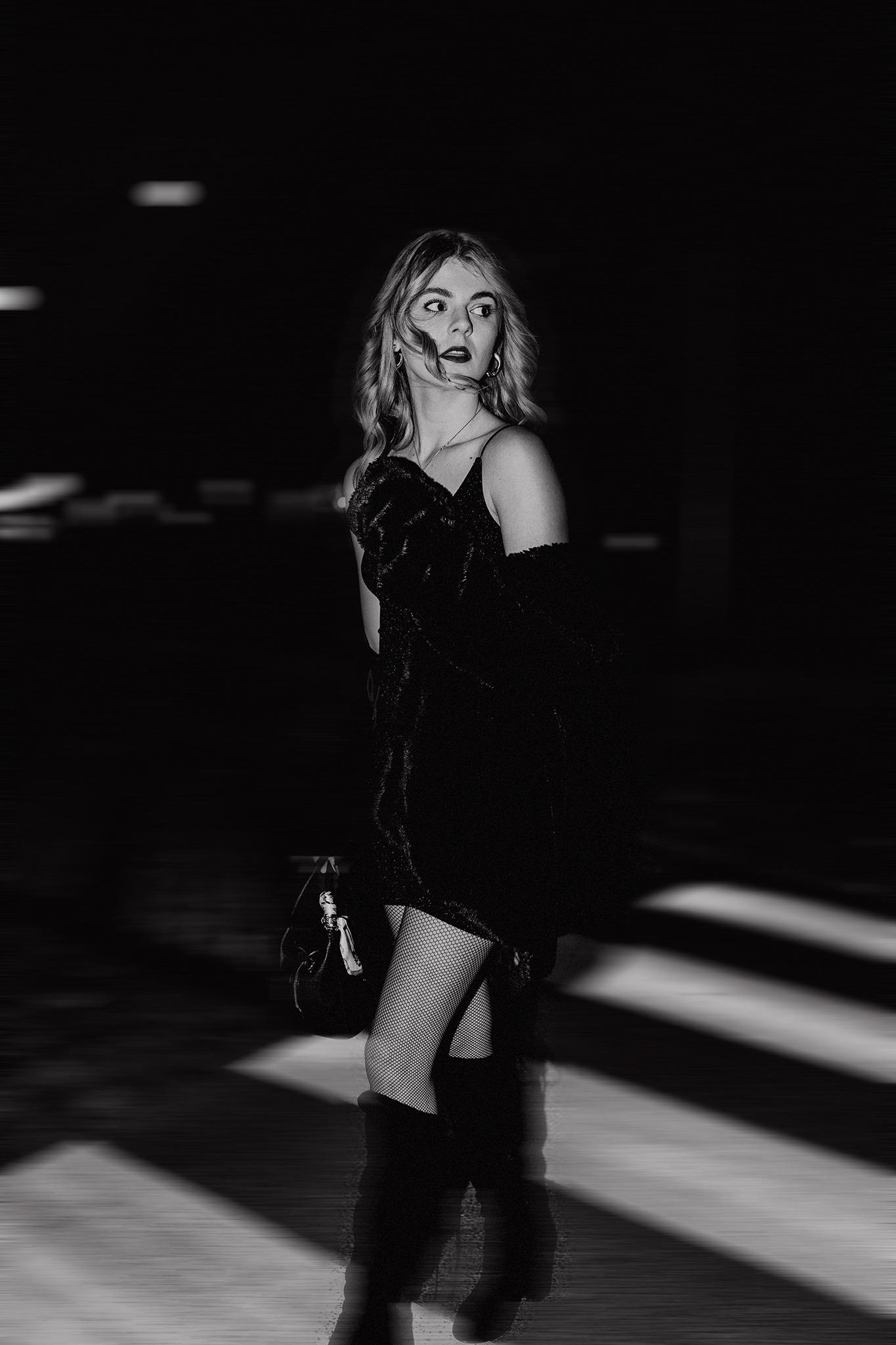 Czarno-białe zdjęcie kobiety w czarnej sukience, kabaretkach i kozakach, z torebką, stojącej na pasach dla pieszych, z dynamicznym rozmyciem tła.