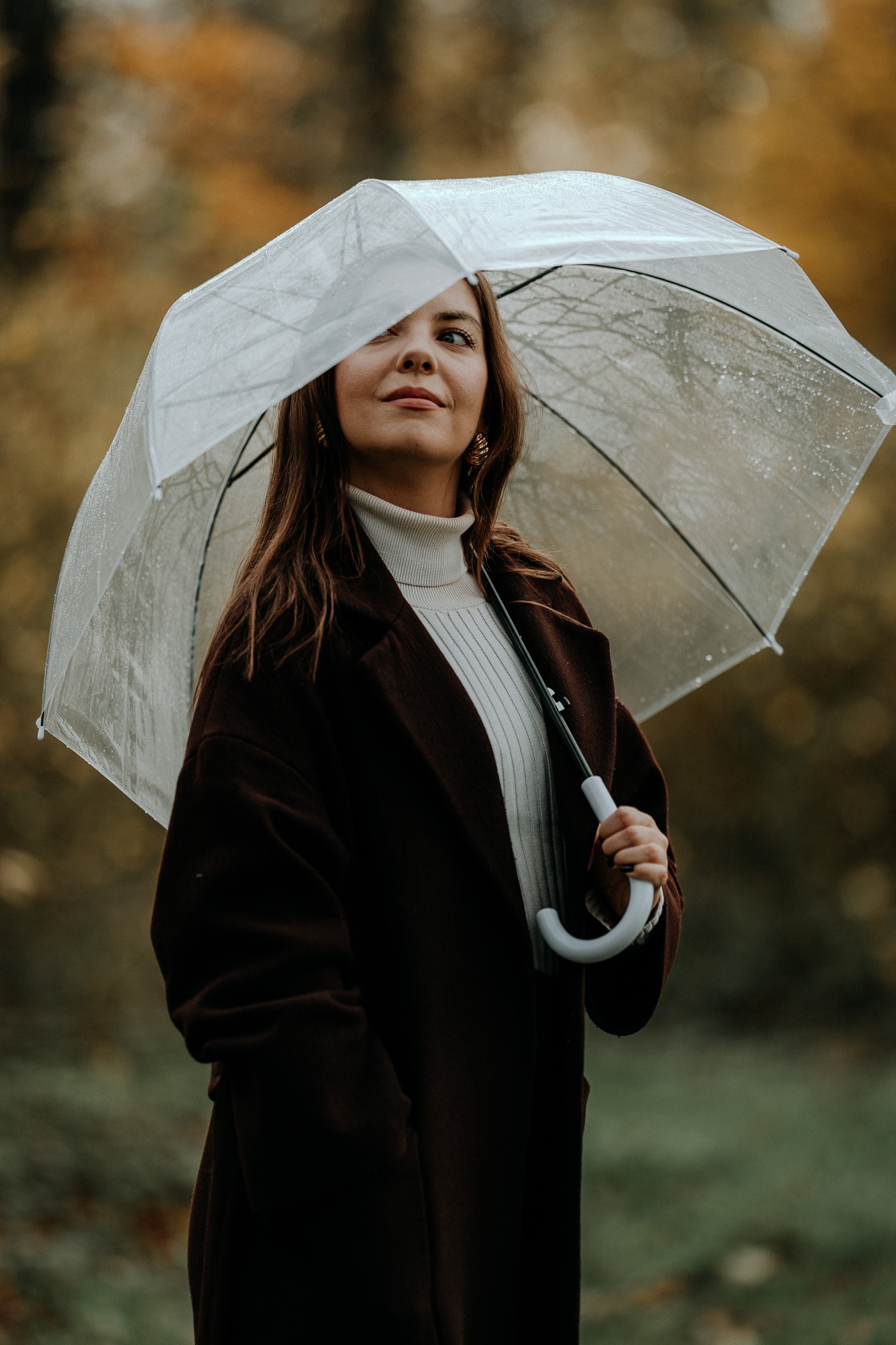 Portret kobiety w brązowym płaszczu i golfie, trzymającej przezroczysty parasol z kroplami deszczu, zamyślona poza w jesiennej scenerii.
