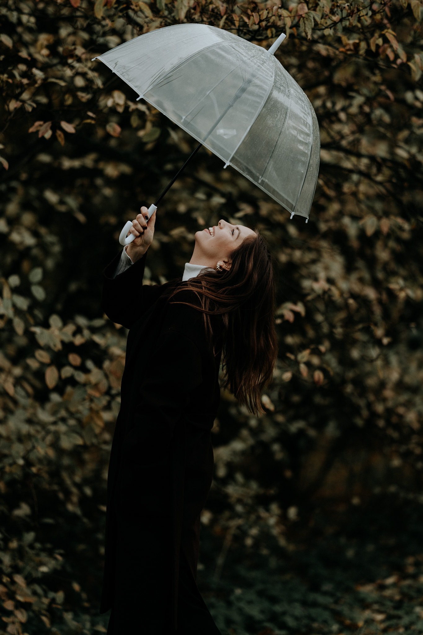 Uśmiechnięta kobieta w czarnym płaszczu trzymająca przezroczysty parasol na tle jesiennych liści.