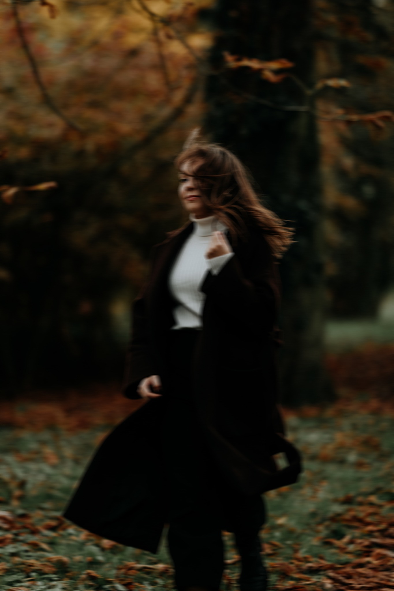 Kobieta w ciemnym płaszczu i białym golfie, włosy rozwiane na wietrze, w tle jesienny park z opadłymi liśćmi, lekko rozmyte ujęcie.