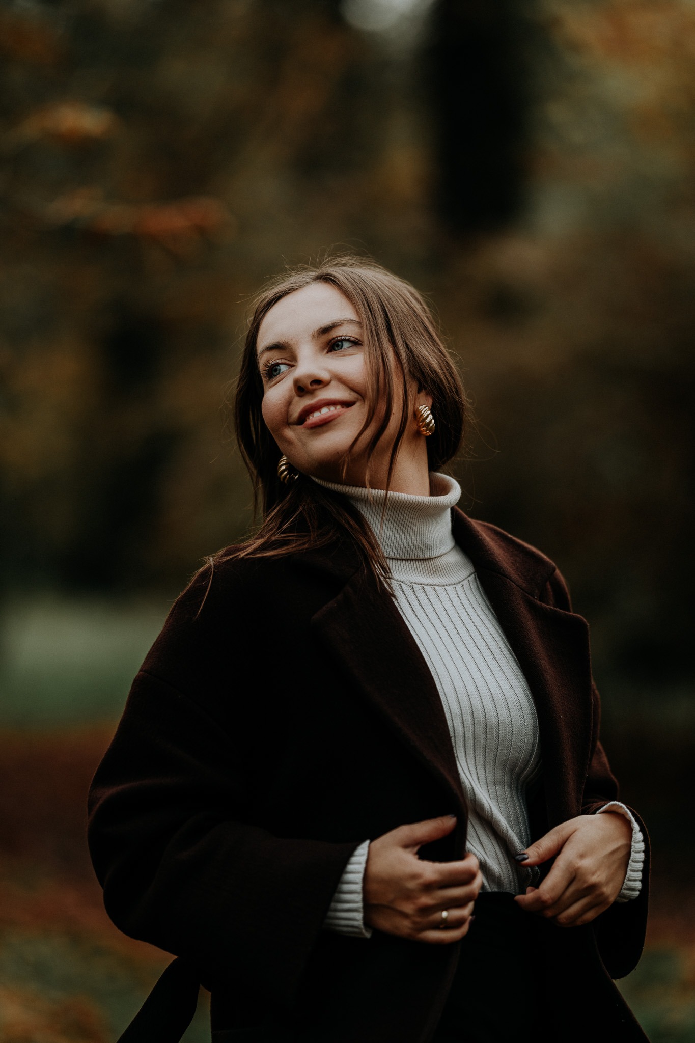 Uśmiechnięta kobieta w brązowym płaszczu i kremowym golfie, pozująca w jesiennym plenerze z rozmytym tłem drzew.