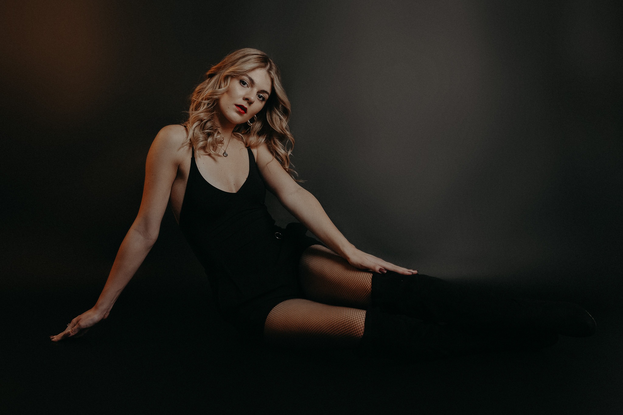 Portret kobiety w czarnym body i pończochach siatkowych, leżącej na ciemnym tle, z delikatnym oświetleniem bocznym podkreślającym jej blond włosy i czerwone usta.
