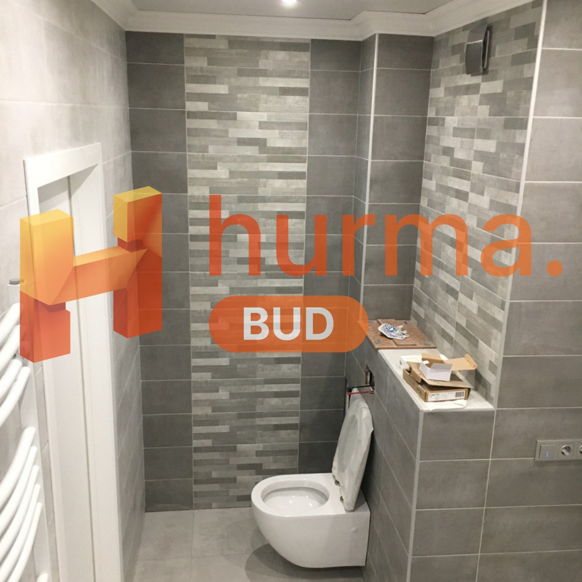 Wykończona łazienka w odcieniach szarości z podwieszaną toaletą, widoczne płytki imitujące cegłę oraz zabudowa geberitu z półką i akcesoriami montażowymi.