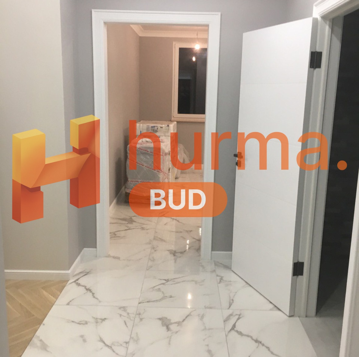 Wykończony korytarz z białymi płytkami imitującymi marmur, otwartymi białymi drzwiami i widocznymi materiałami budowlanymi w sąsiednim pomieszczeniu, z logo firmy nałożonym na zdjęcie.