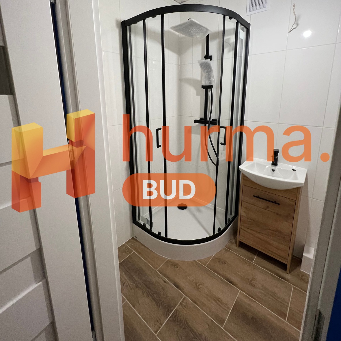 Nowoczesna kabina prysznicowa z czarnymi profilami, umywalka z szafką w odcieniach drewna, białe płytki i podłoga imitująca drewno, widoczne logo firmy.