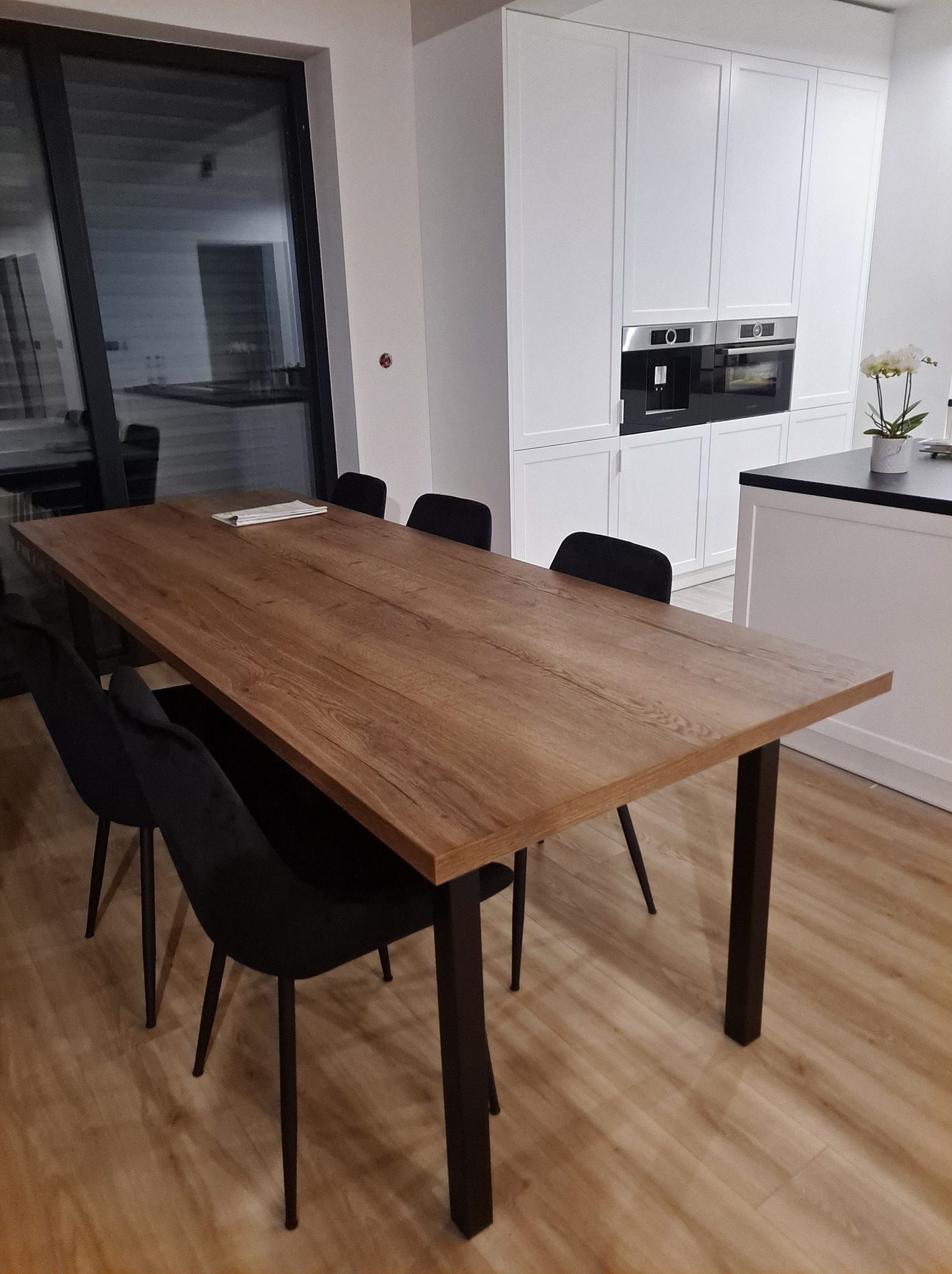 Stół z blatem drewnopodobnym i czarnymi, metalowymi nogami w nowoczesnej jadalni z białymi szafkami kuchennymi w tle.