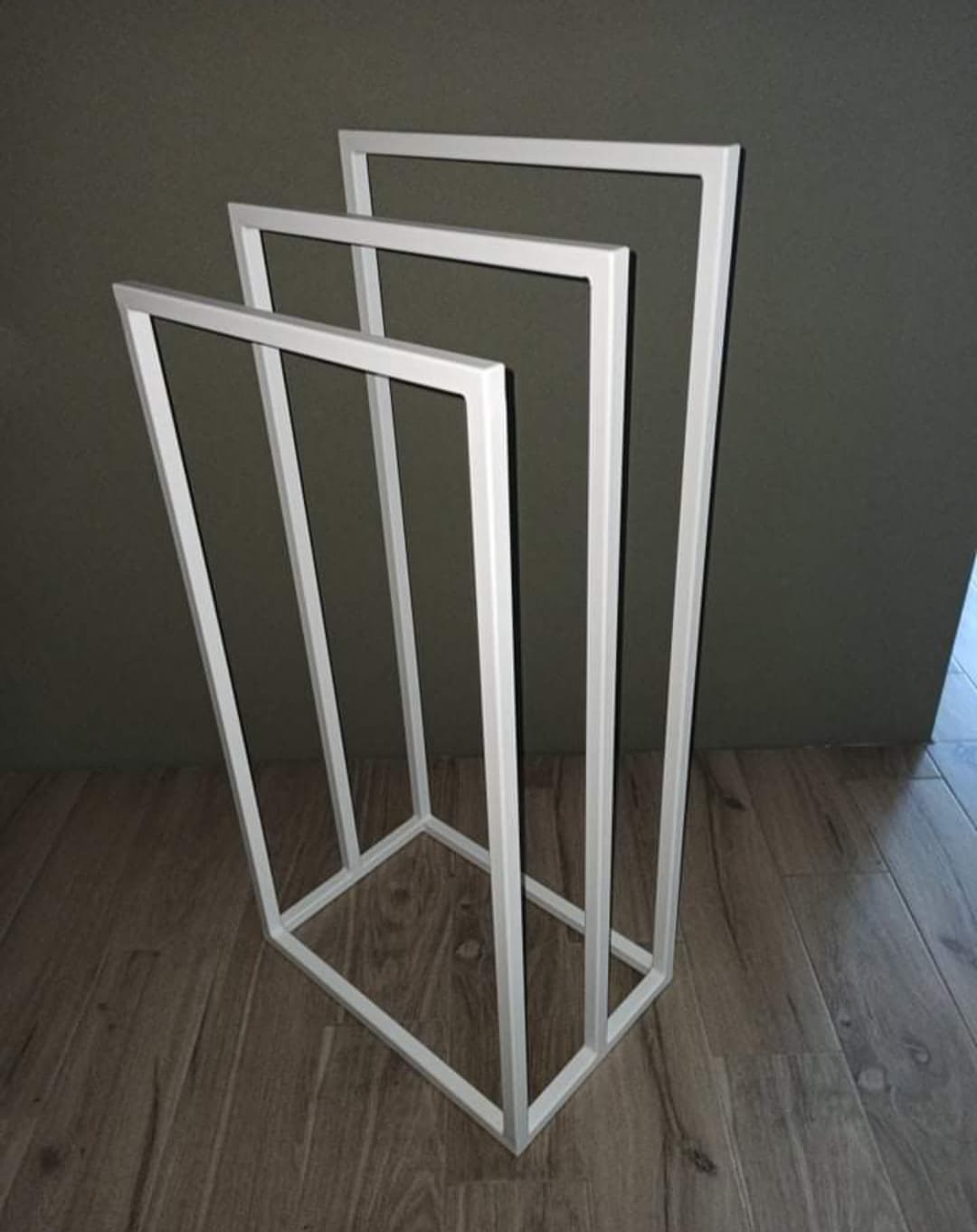 Biały, minimalistyczny, metalowy stojak na ręczniki, o geometrycznej konstrukcji, na drewnianej podłodze, na tle ciemnej ściany.