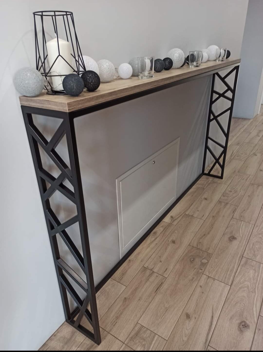 Konsola z drewnianym blatem i metalową podstawą w geometryczne wzory, udekorowana świecą, szklankami i kulami dekoracyjnymi, na tle drewnianej podłogi