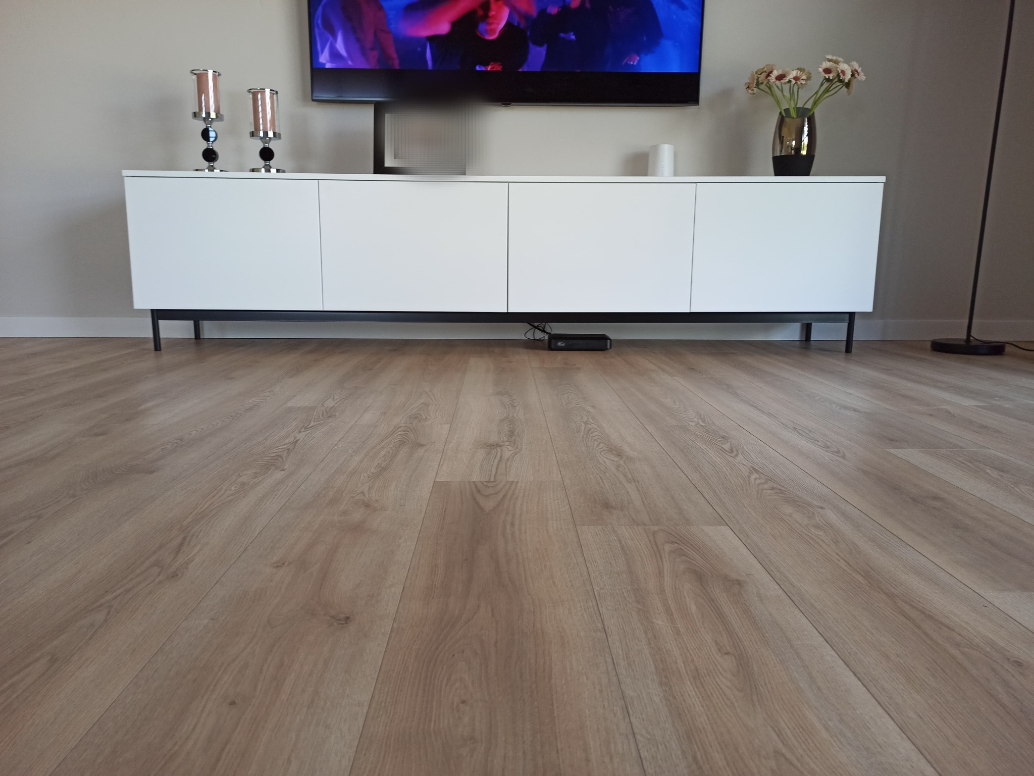 Jasnobrązowa podłoga laminowana z widoczną strukturą drewna, biała szafka RTV z czarnymi nogami, telewizor na ścianie oraz dekoracje.