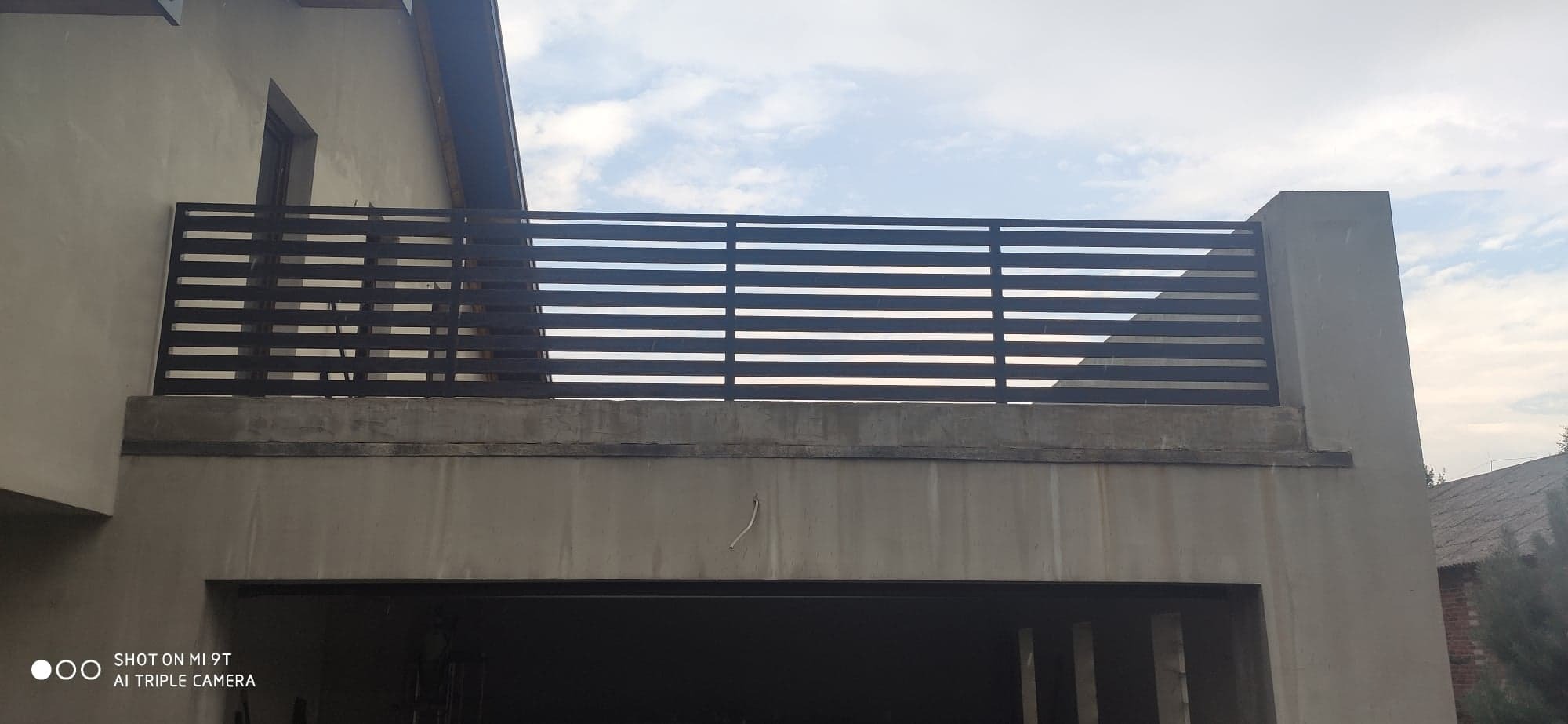 Nowoczesna, pozioma balustrada z metalowych profili na betonowym balkonie, widok z dołu na tle pochmurnego nieba.