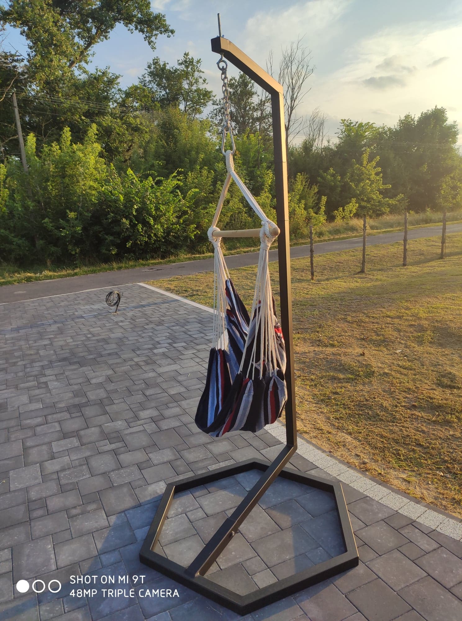 Metalowy stelaż z wiszącym hamakiem w paski na kostce brukowej, w tle zieleń i droga.