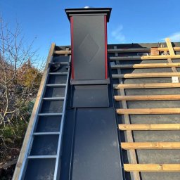 Czar-Bud - Dach w trakcie budowy w Filipowie: czarna blacha na łatach, komin z elementami czerwieni i drabina oparta o dach. Widok z dołu na błękitne niebo.