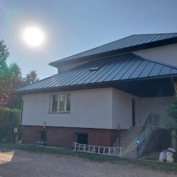 Czar-Bud - Dom jednorodzinny z ciemnym, metalowym dachem i nową elewacją w jasnym odcieniu. Widoczne rynny, okno dachowe i schody wejściowe. Słoneczny dzień.