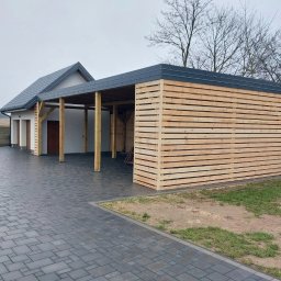 Czar-Bud - Nowoczesna wiata garażowa z drewnianą ażurową ścianą, przylegająca do budynku mieszkalnego z szarym dachem. Uwagę przykuwa staranne wykonanie i estetyczne połączenie materiałów.