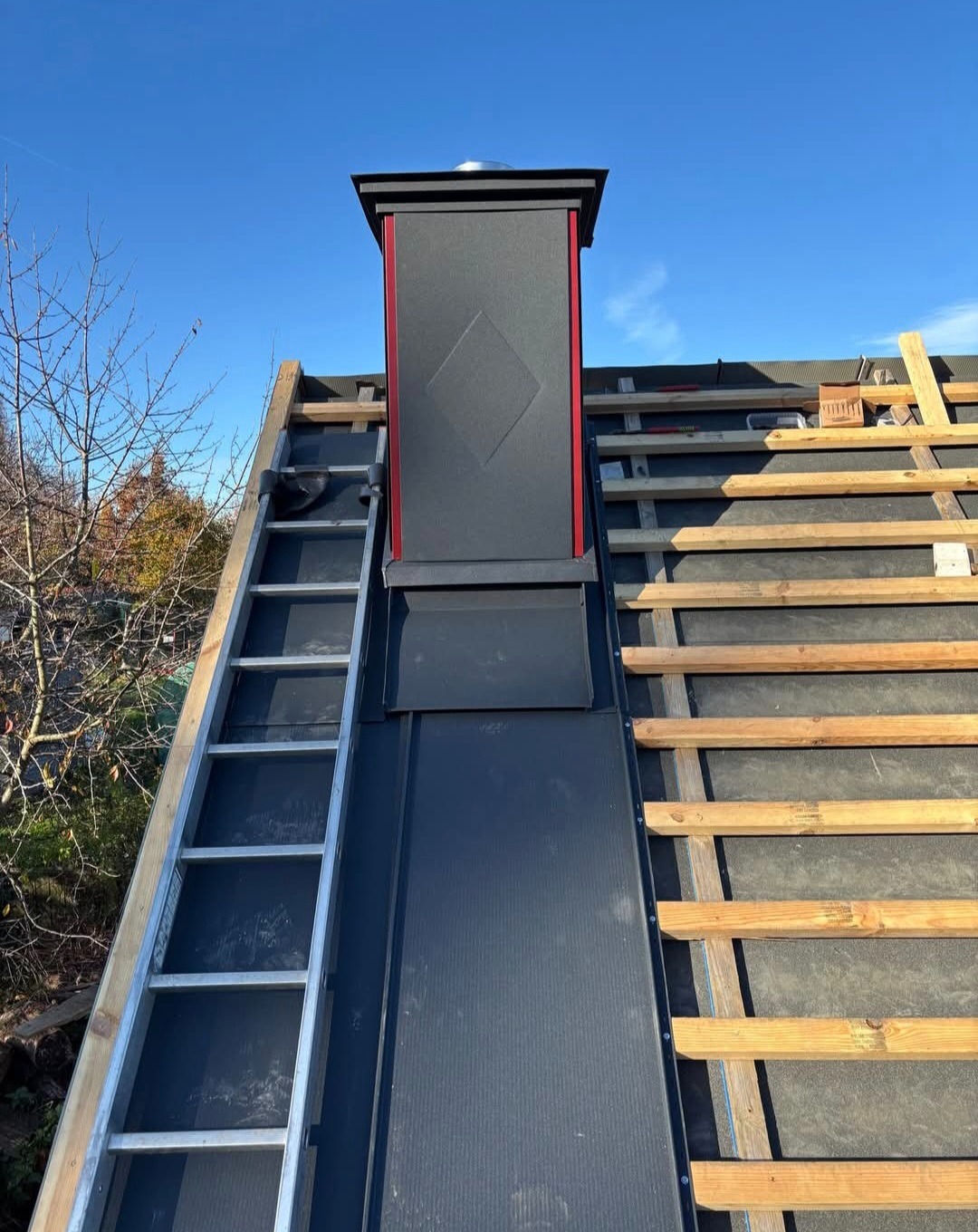 Dach w trakcie budowy w Filipowie: czarna blacha na łatach, komin z elementami czerwieni i drabina oparta o dach. Widok z dołu na błękitne niebo.