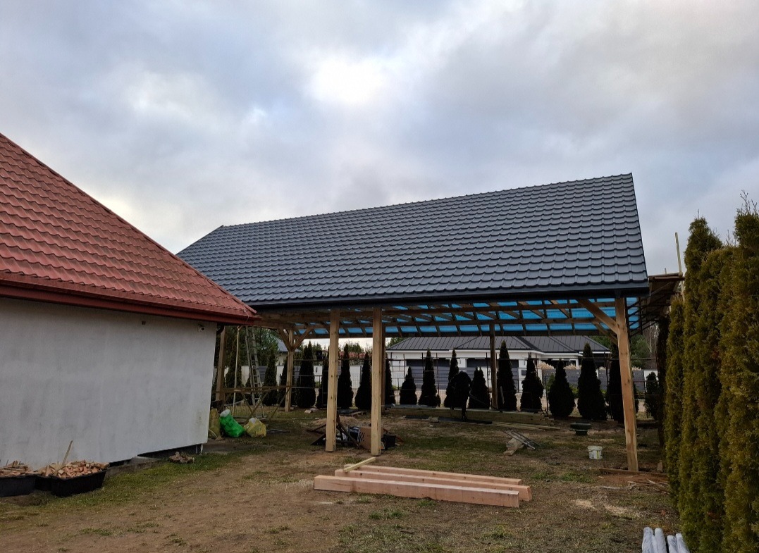 Zadaszenie tarasu z ciemnoszarą dachówką, widoczne drewniane słupy konstrukcyjne i fragment dachu budynku z czerwoną dachówką. Tło: pochmurne niebo.