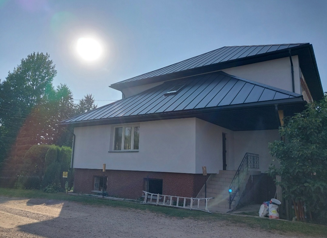 Dom jednorodzinny z ciemnym, metalowym dachem i nową elewacją w jasnym odcieniu. Widoczne rynny, okno dachowe i schody wejściowe. Słoneczny dzień.