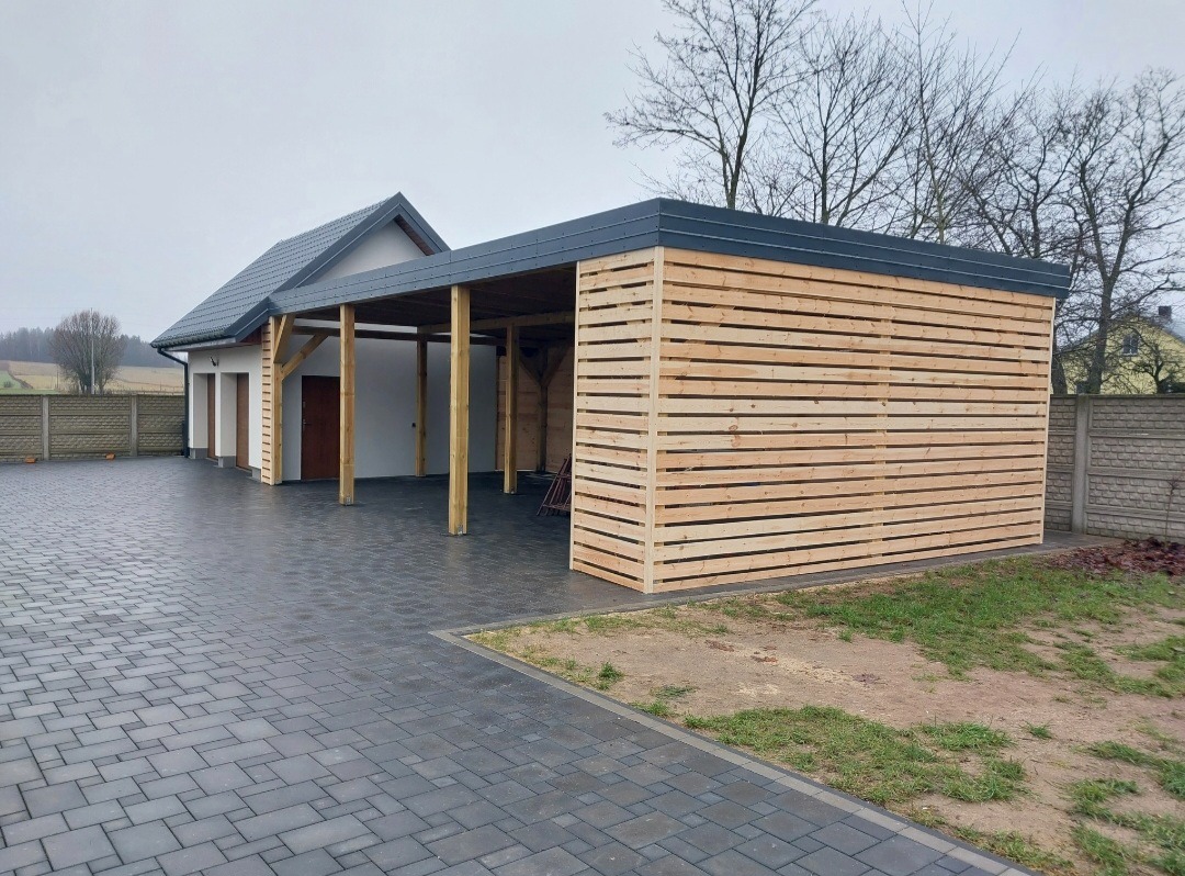 Nowoczesna wiata garażowa z drewnianą ażurową ścianą, przylegająca do budynku mieszkalnego z szarym dachem. Uwagę przykuwa staranne wykonanie i estetyczne połączenie materiałów.