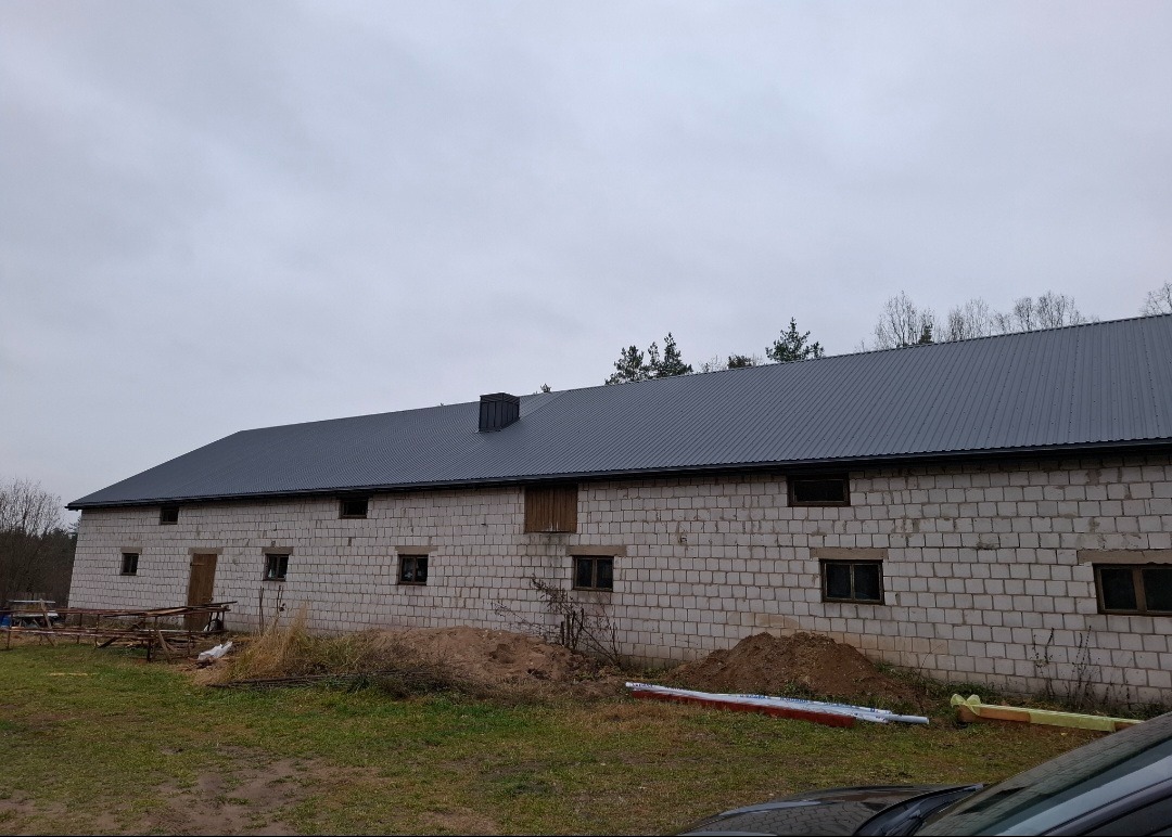 Budynek gospodarczy z szarego bloczka, z nowym, ciemnoszarym dachem z blachy trapezowej. Widoczne ślady prac budowlanych na trawniku przed budynkiem.