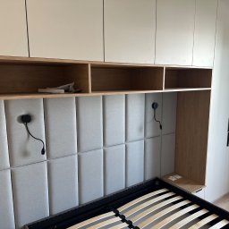 BartServices Bartłomiej Leśniak - Nowoczesna sypialnia z zabudową meblową: białe szafy, drewniane półki nad wezgłowiem łóżka z tapicerowanym panelem ściennym i regulowanymi lampkami nocnymi.