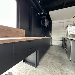 BartServices Bartłomiej Leśniak - Nowoczesna zabudowa kuchenna z czarnymi frontami i drewnianym blatem w lokalu gastronomicznym, widoczne elementy wentylacji sufitowej i chłodnicze witryny.