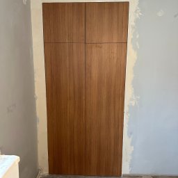 BartServices Bartłomiej Leśniak - Montaż czterodrzwiowej szafy wnękowej z okładziną drewnopodobną na tle przygotowywanej ściany, widoczny poziomica i materiały montażowe.
