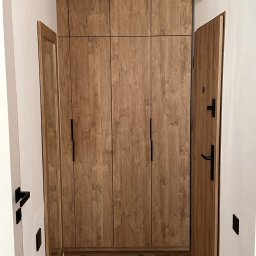 BartServices Bartłomiej Leśniak - Wysoka, drewniana szafa z czarnymi uchwytami, zabudowana od podłogi do sufitu w wąskim korytarzu, z widocznym lustrem w drewnianej ramie i otwartymi drzwiami wejściowymi.