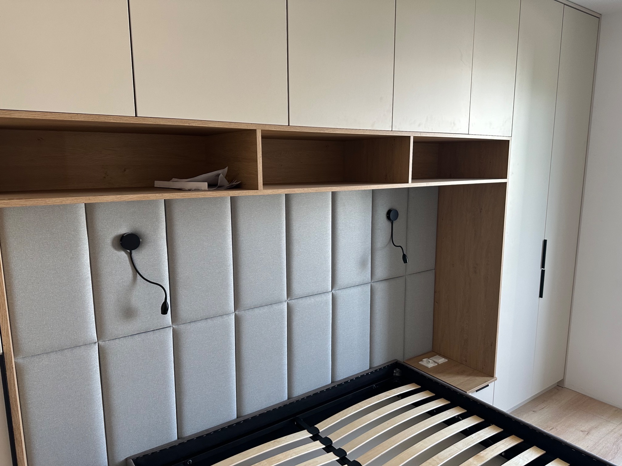 Nowoczesna sypialnia z zabudową meblową: białe szafy, drewniane półki nad wezgłowiem łóżka z tapicerowanym panelem ściennym i regulowanymi lampkami nocnymi.