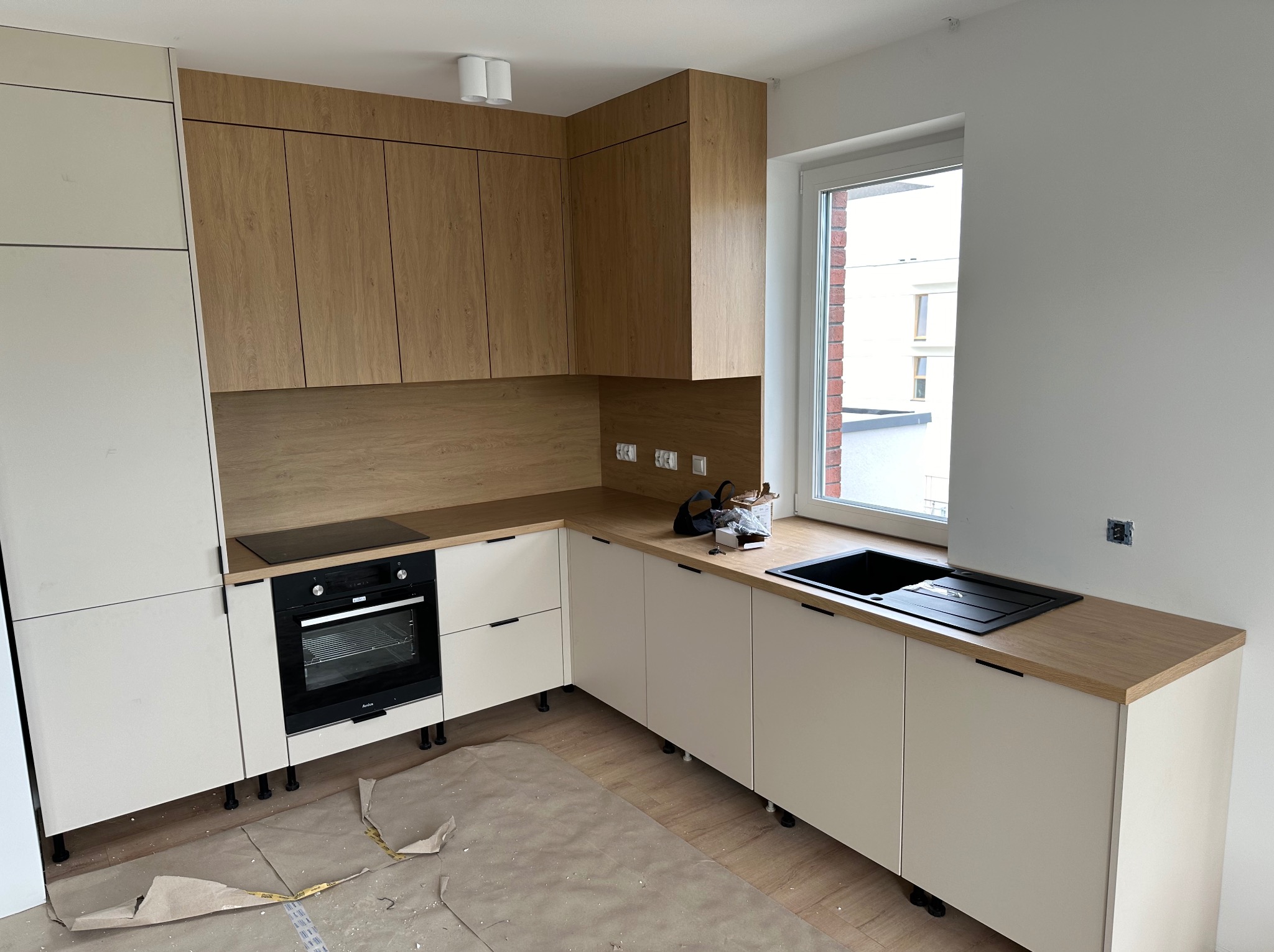 Nowoczesna kuchnia z jasnymi, drewnopodobnymi szafkami górnymi i kremowymi dolnymi, czarnym zlewem i płytą grzewczą, widoczne ślady montażu na podłodze.