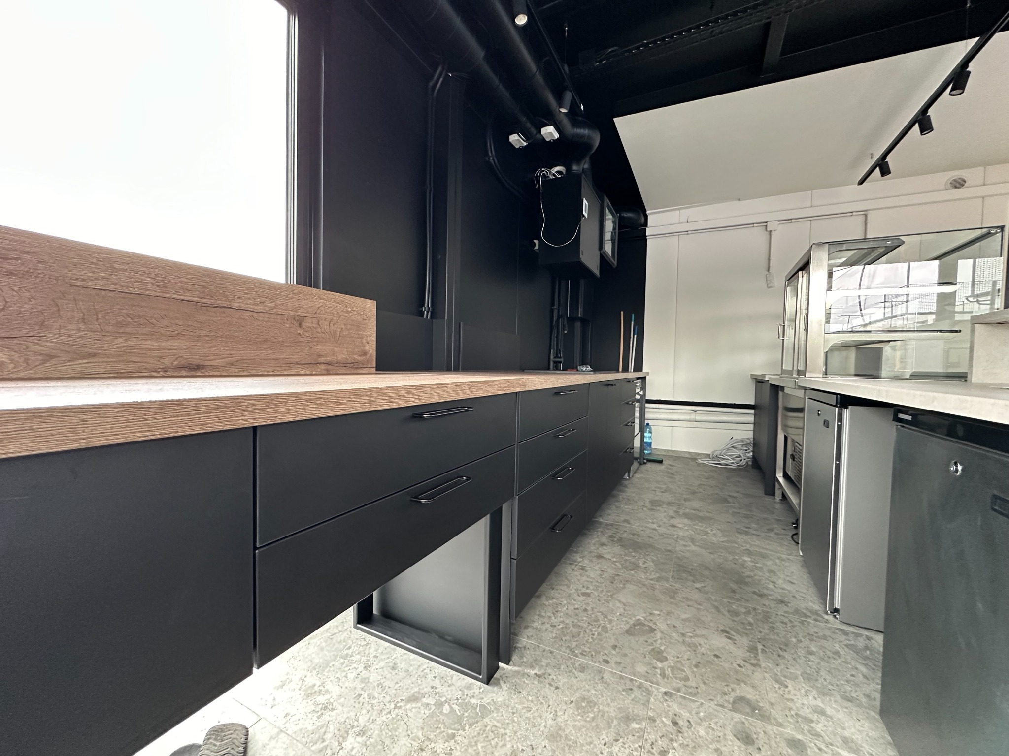 Nowoczesna zabudowa kuchenna z czarnymi frontami i drewnianym blatem w lokalu gastronomicznym, widoczne elementy wentylacji sufitowej i chłodnicze witryny.