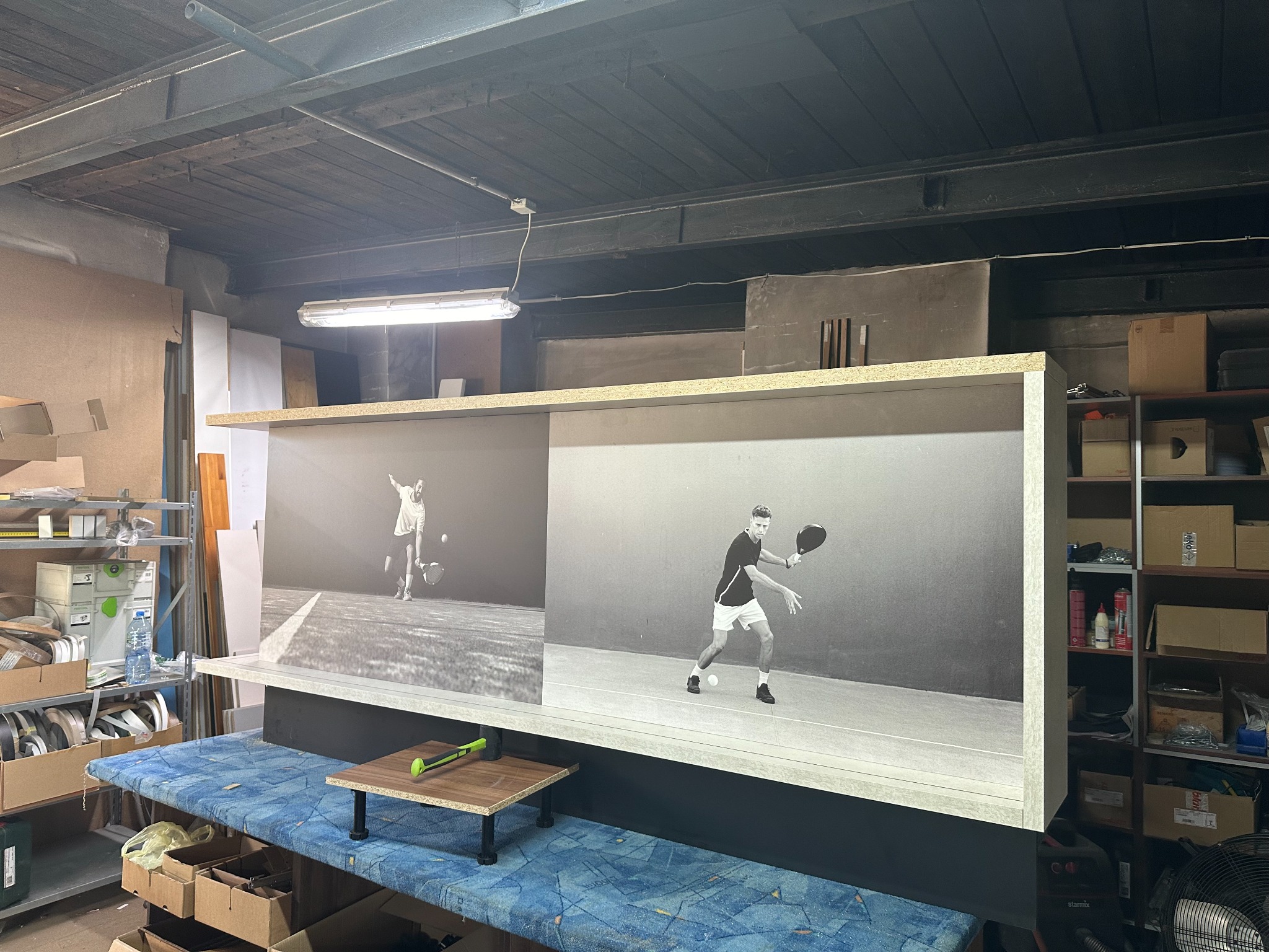 Niedokończona szafa z nadrukowanymi czarno-białymi zdjęciami tenisistów, stojąca w warsztacie z widocznym narzędziem i niebieskim blatem roboczym.