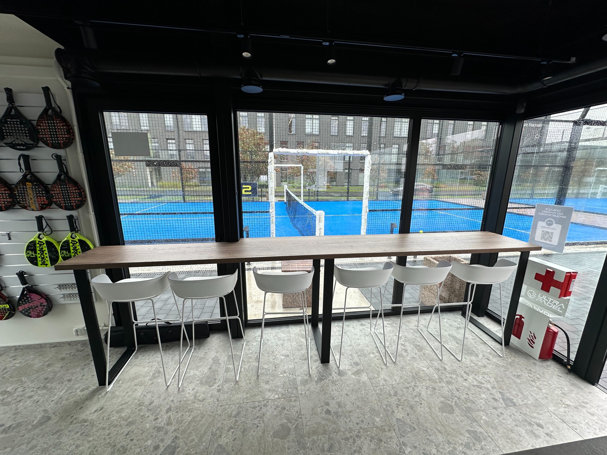 Wnętrze lokalu z widokiem na kort do padla: drewniany blat baru z białymi hokerami, ściana z rakietami do padla, apteczka pierwszej pomocy.