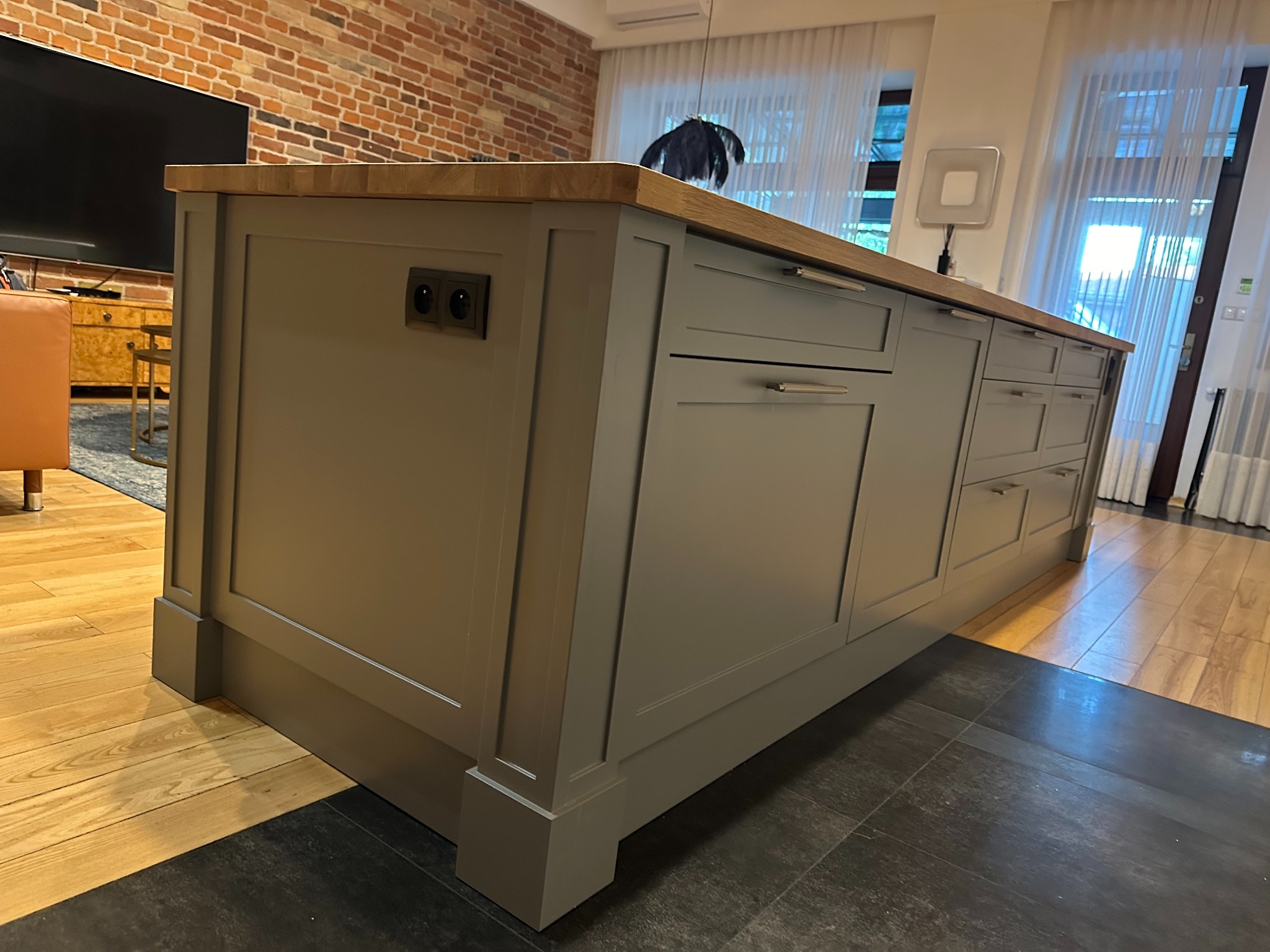 Szara wyspa kuchenna z drewnianym blatem, widoczne gniazdka elektryczne, na tle ceglanej ściany i zasłon w oknie.