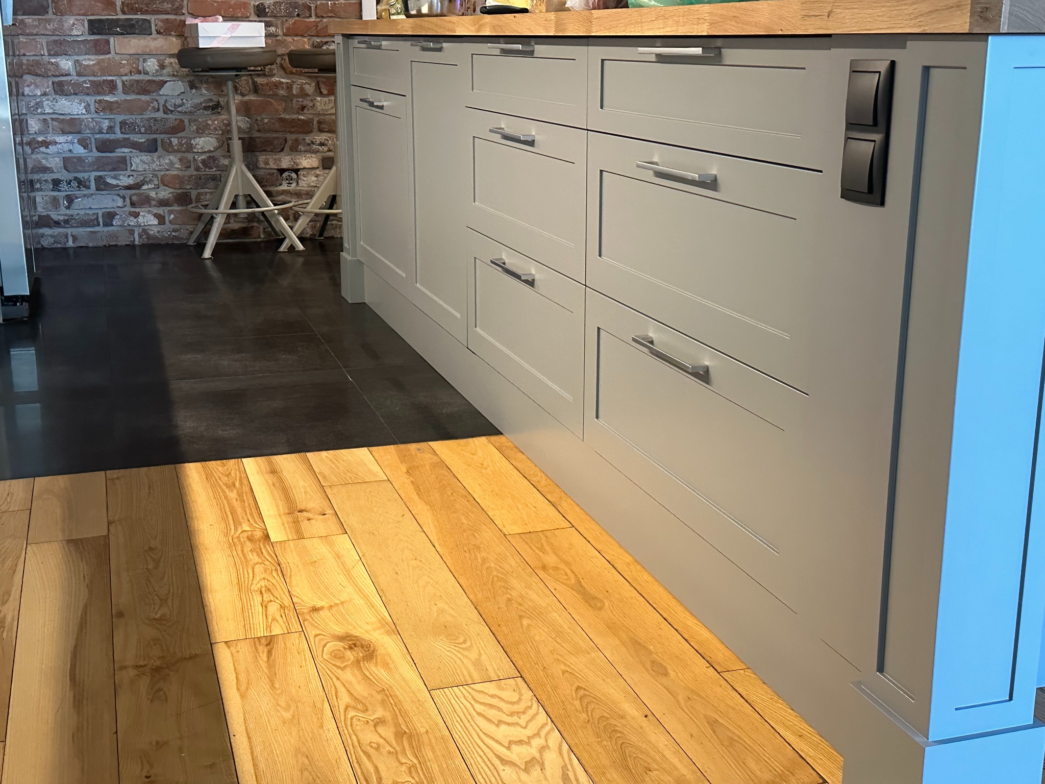 Szara szafa na wymiar z drewnianym blatem, metalowymi uchwytami i czarnymi włącznikami światła, widoczna z perspektywy podłogi z drewnianymi panelami w ciepłym świetle.