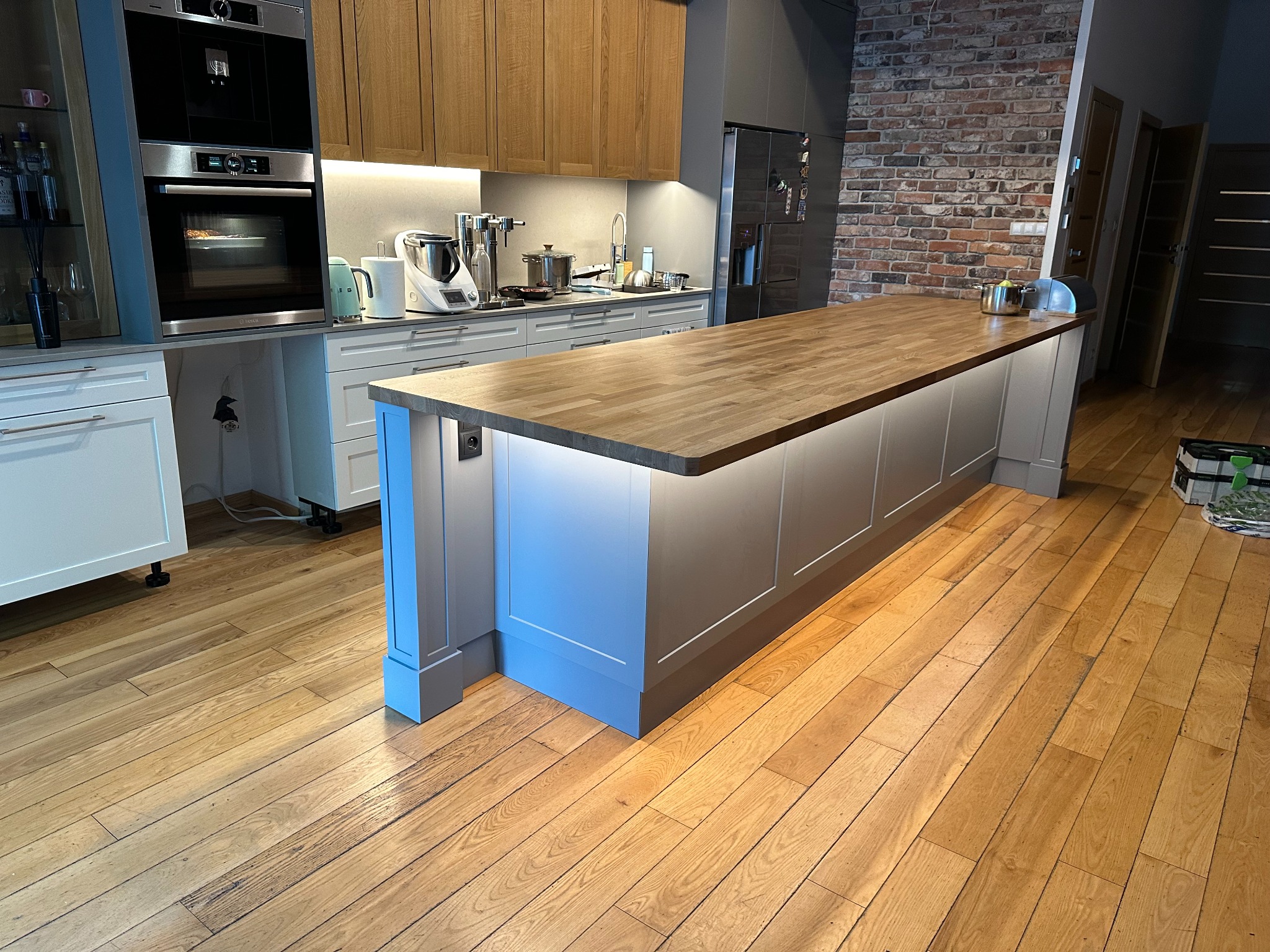 Nowoczesna kuchnia z wyspą o drewnianym blacie, szare szafki z subtelnym oświetleniem LED, drewniana podłoga i ściana z cegły w tle.