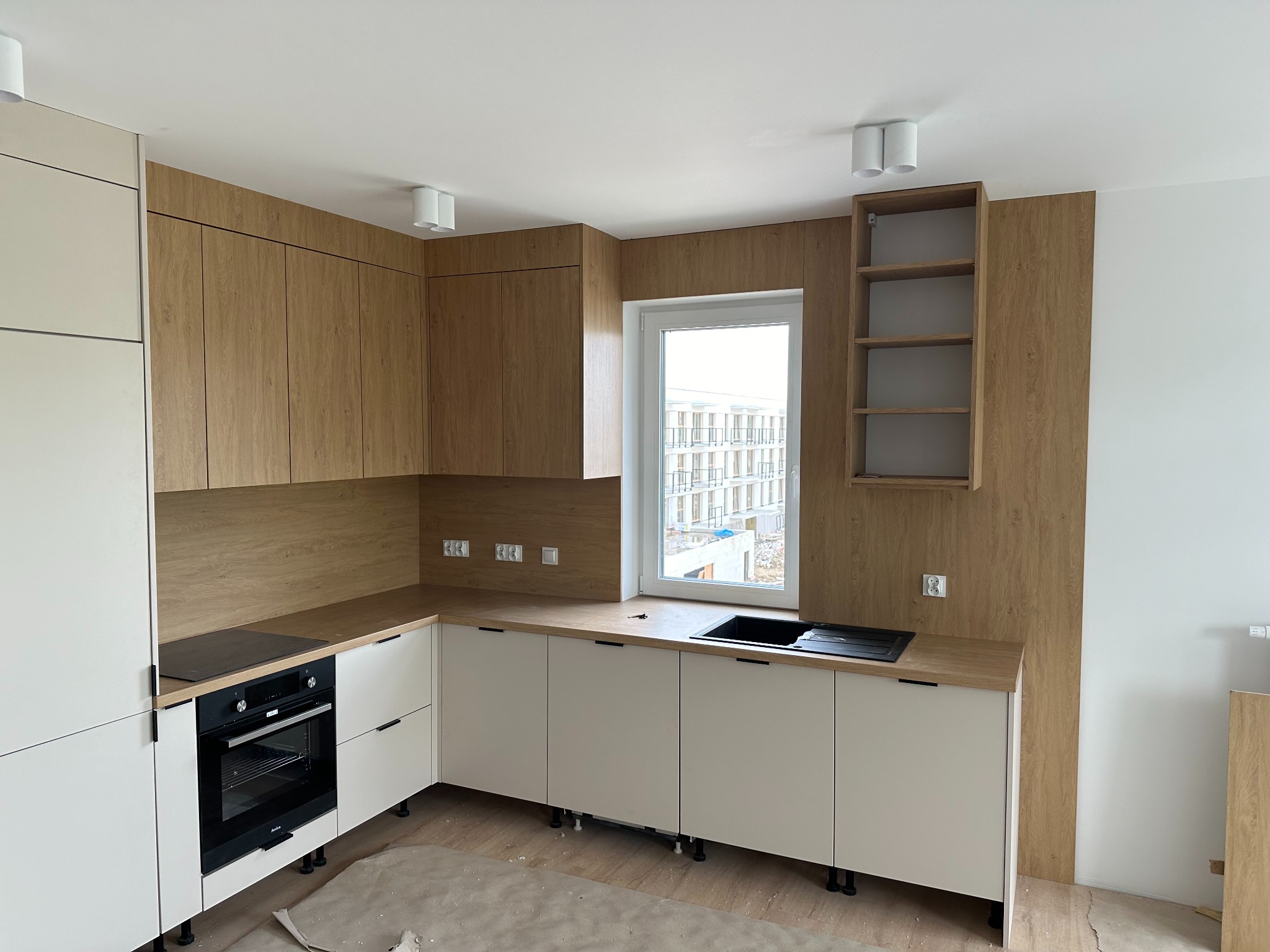 Nowoczesna kuchnia w trakcie montażu, z białymi szafkami dolnymi i drewnopodobnymi górnymi, widoczne elementy zabudowy piekarnika i płyty grzewczej, okno z widokiem na budynek w budowie, podłoga...