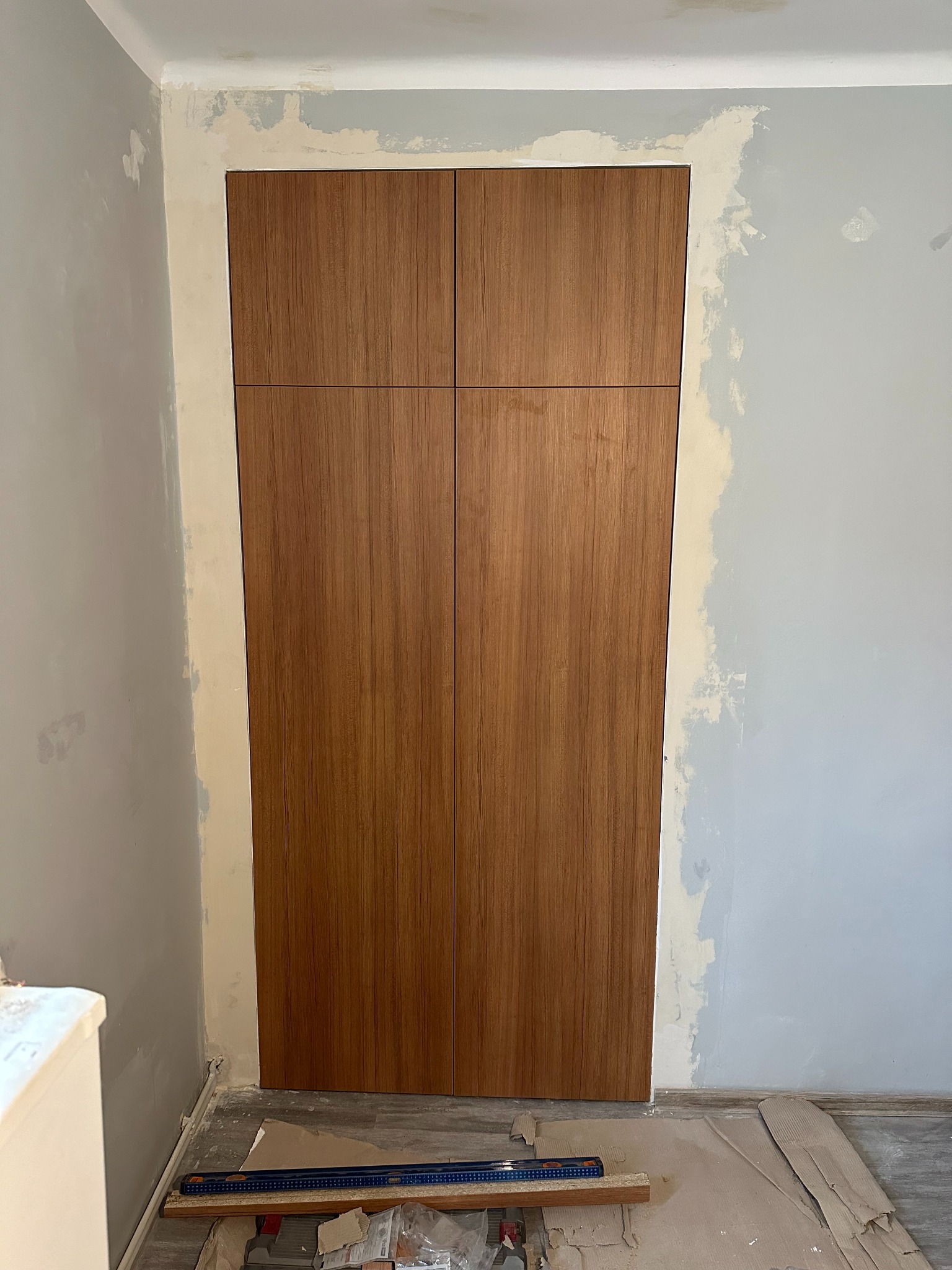 Montaż czterodrzwiowej szafy wnękowej z okładziną drewnopodobną na tle przygotowywanej ściany, widoczny poziomica i materiały montażowe.