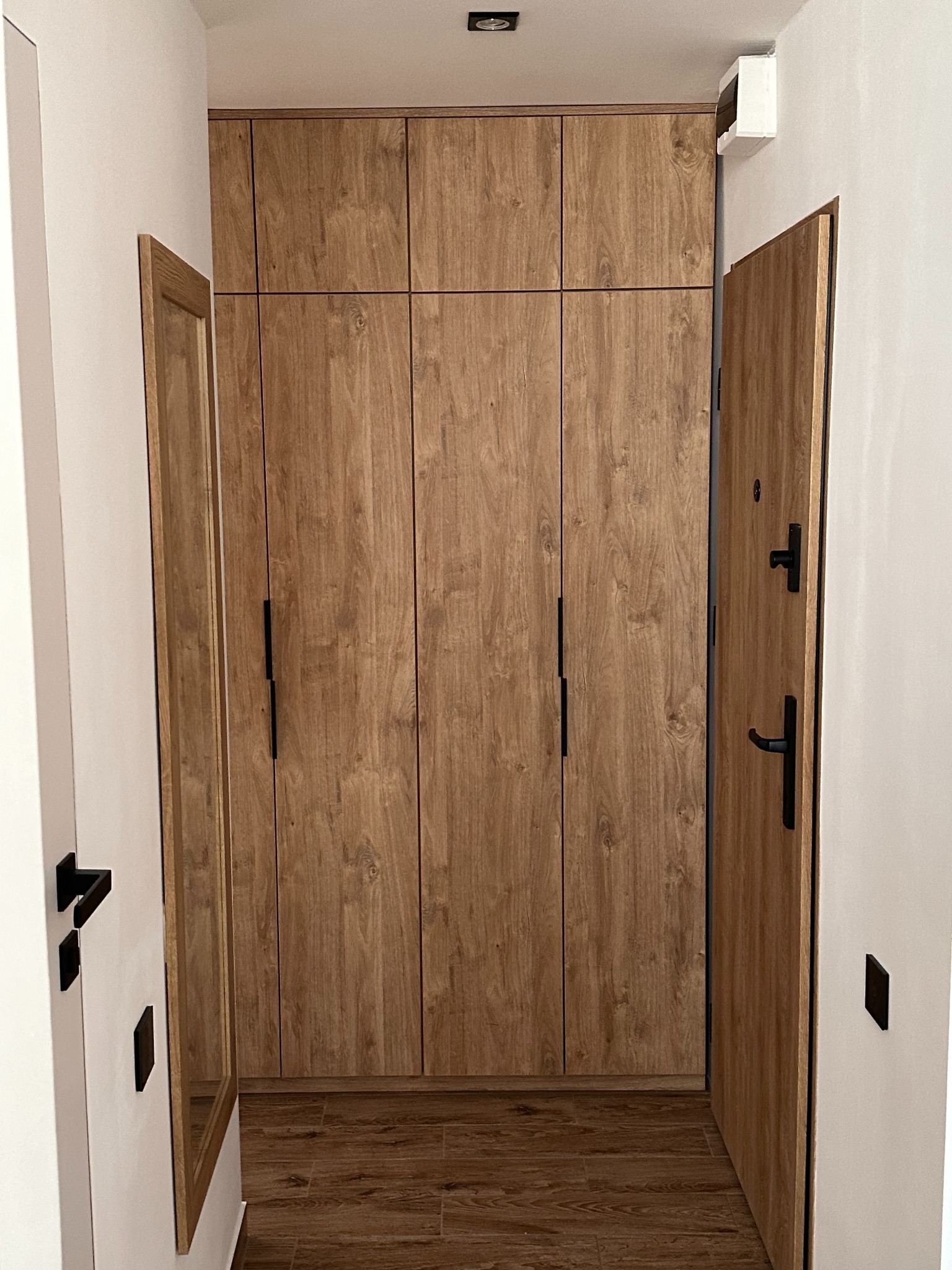 Wysoka, drewniana szafa z czarnymi uchwytami, zabudowana od podłogi do sufitu w wąskim korytarzu, z widocznym lustrem w drewnianej ramie i otwartymi drzwiami wejściowymi.