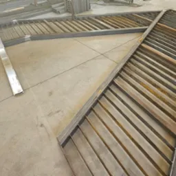 Metalowa konstrukcja schodów z kątowników i prętów, częściowo zardzewiała, leżąca na betonowej posadzce, widoczne narzędzia ślusarskie w tle.