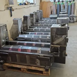 Metalowe elementy konstrukcyjne na paletach w warsztacie, widoczne ślady spawania, butle z gazem w tle.